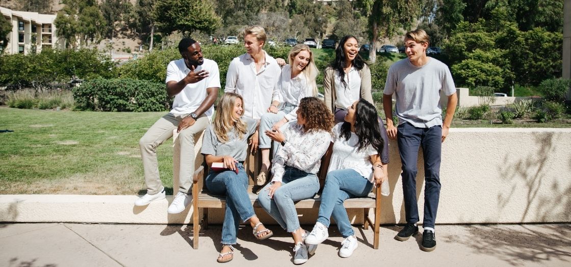 A group of students sit on and above a bench talking and laughing